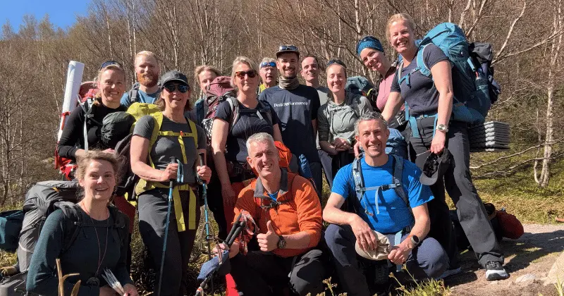 Een groep wandelaars met rugzakken en uitrusting lacht voor een foto in de buitenlucht op een zonnige dag, omringd door kale bomen en groen.